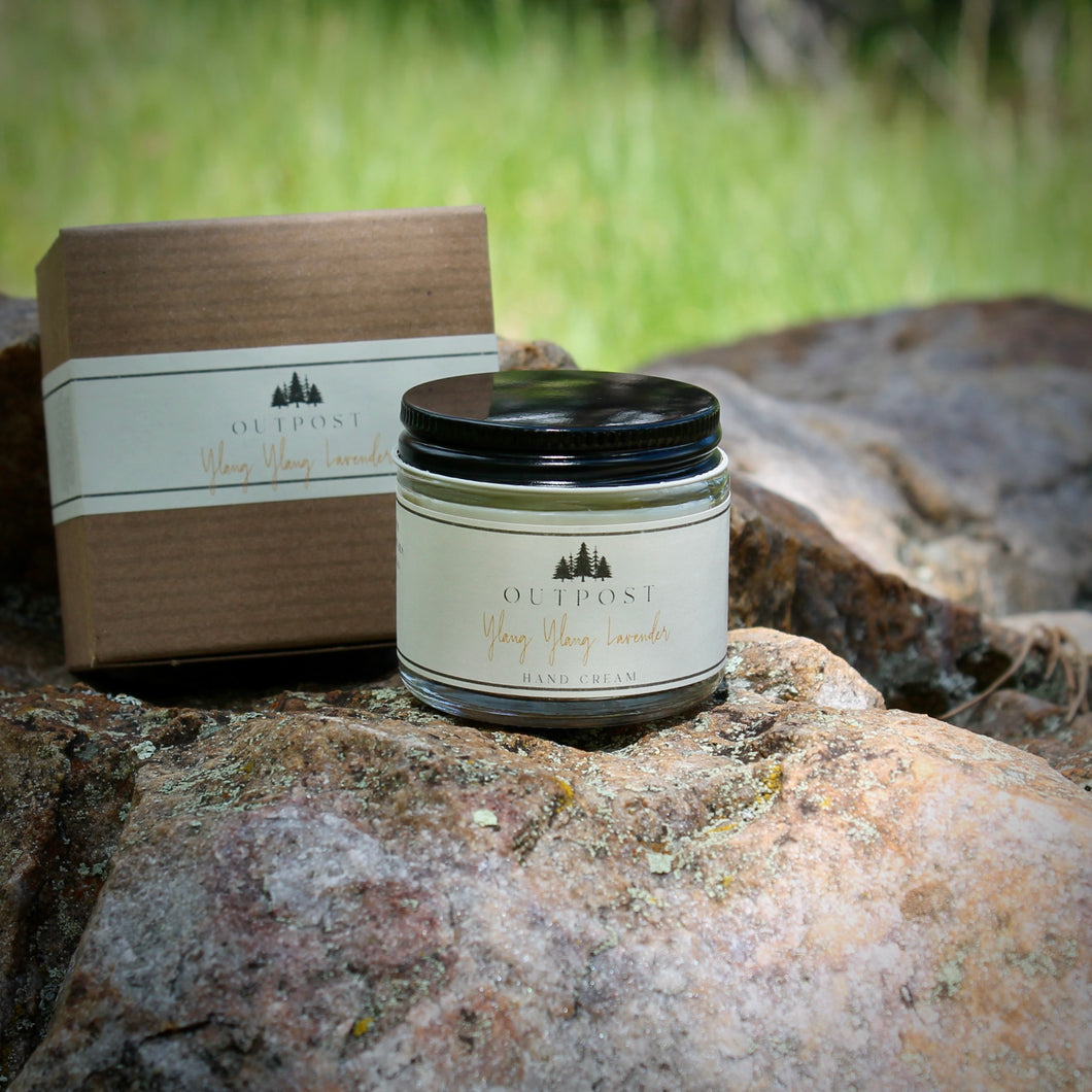 a glass jar of Ylang Ylang Lavender hand cream sits on a large boulder in dappled shade with a kraft gift box behind sitting behind 